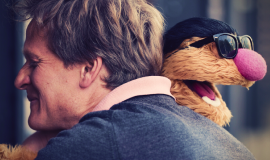 Photograph of a man hugging an orange puppet wearing sunglasses